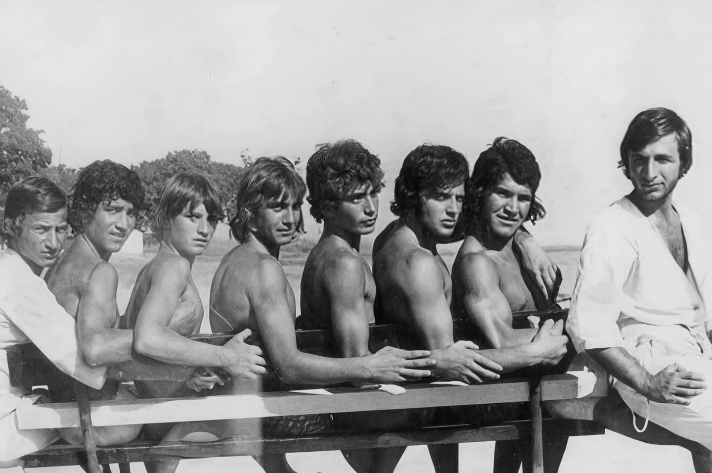 A black and white photo of 8 sun-tanned young men sitting on the bench with natural landscape behind.